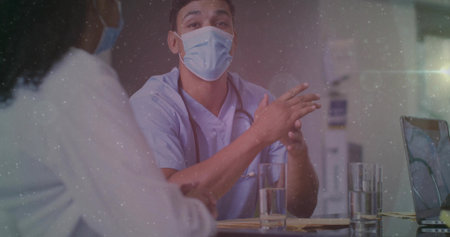 Speaking doctor in scrubs and face mask at clinic desk, with stethoscope laptop papers water. Healthcare, medical, professional, consultation, communication, diagnosis, wellnessの写真素材