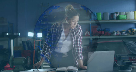 Leaning Asian woman mechanic wearing plaid shirt, jeans, checking laptop in garage, with car engine. Industrial, craftsmanship, workspace, technical, hands-on, engineering, toolsの写真素材