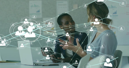 Two businesswomen in blazers collaborating using laptops and tablet in office, with network overlay. Professional, teamwork, innovation, communication, digital, analytics, workplaceの写真素材