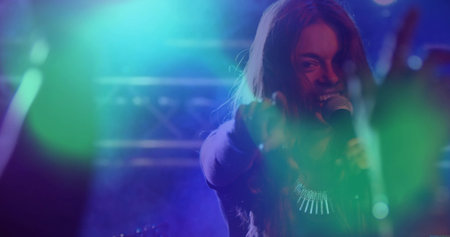 Performing singer wearing necklace holding microphone on stage under spotlights, with blurred hands....