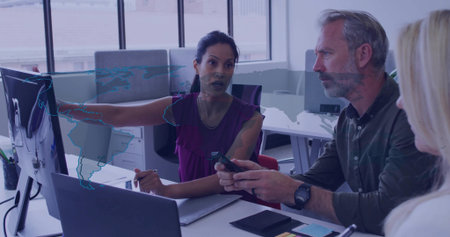 Gesturing dark-haired woman pointing at computer monitor in office, with laptop and smartphone....