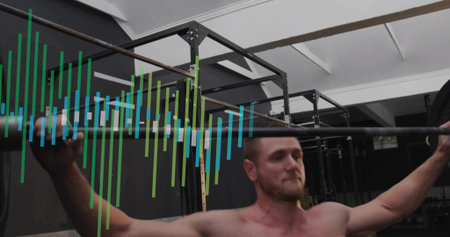 Lifting shirtless man pressing barbell overhead in gym weight area with pullup rig and colored bars....