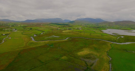 Meandering stream flowing through green fields in valley, with stone walls hedgerows and farmhouses....