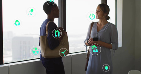 Two colleagues discussing sustainability ideas at office windows with smartphone and eco-icons....