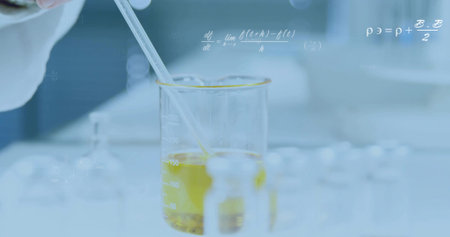 Gloved hand dispensing yellow solution with pipette into beaker at lab bench, with vials, flasks. Science, experimentation, chemistry, research, laboratory, innovation, precisionの写真素材