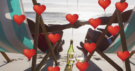 Swimwear-clad couple holding hands on chairs by ocean with wine bottle, red balloons. Romantic, leisure, destination, vacation, serenity, lifestyle, relaxationの写真素材