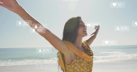 Standing woman wearing yellow sundress raising arms at beach, with floating social media icons. Joy,...