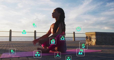 Meditating woman in sportswear sitting on pink mat at seaside promenade, with floating energy icons. Wellness, sustainability, renewable, meditation, outdoor, tranquility, environmentの写真素材