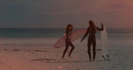 Holding pink and white surfboards, couple facing each other on sandy beach, full-length wetsuits....
