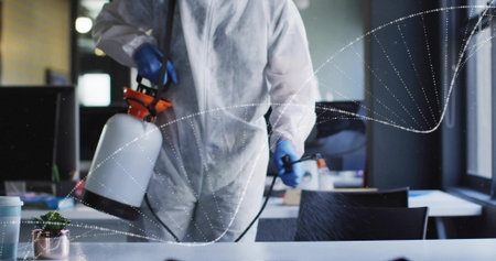 Spraying cleaning worker wearing protective suit disinfecting office desk, with sprayer and plant. Disinfection, sanitation, safety, hygiene, modern, professional, sterileの写真素材
