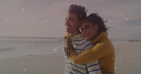 Hugging couple smiling, gazing at horizon on beach at twilight, with mustard and navy sweaters. Romance, intimacy, serenity, sunset, leisure, harmony, connectionの写真素材