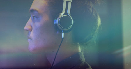 Capturing adult Asian male wearing silver headphones in studio, with blue-green bokeh lighting....