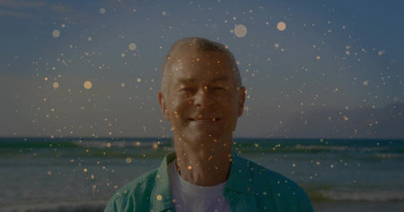 Standing senior man wearing turquoise shirt and white tee at sandy beach, with golden bokeh overlay....