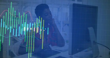 Gazing office worker wearing red blouse resting chin on hand at desk with computer monitor, charts....