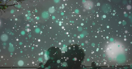 Sitting couple wearing winter coats admiring snow at nighttime park bench, with railing behind....