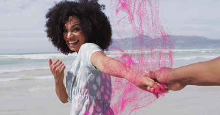 Smiling woman wearing dress holding partner hand pulling along beach with pink powder, copy space....