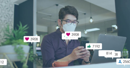 Masked man holding smartphone and viewing social media icons in office with laptop, coffee mug....