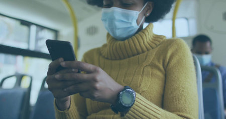 Commuter wearing surgical mask and mustard sweater using smartphone on bus with watch and handrails....