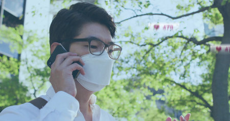 Talking Asian man holding smartphone amid trees near office front, wearing face mask, eyeglasses. Urban, professional, modern, foliage, outdoor, communication, lifestyleの写真素材