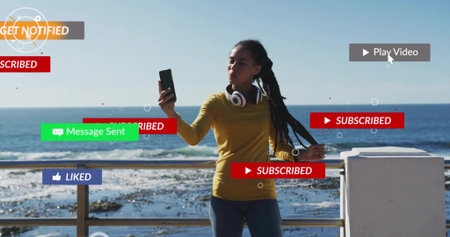 Holding smartphone while wearing headphones on seaside promenade by metal railing, with floating UI....