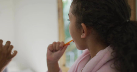 Brushing daughter in bathrobe using orange brush at sink with mirror, mom holding brush, copy space. Family, hygiene, routine, warmth, innocence, domestic, caretakingの写真素材
