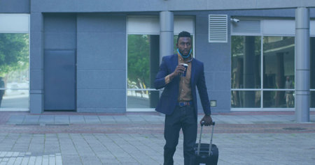 Striding man wearing navy blazer across office plaza pulling suitcase while holding coffee cup....