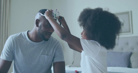 Daughter placing plastic crown on father sitting on bed in bedroom, with tufted headboard. Family, bonding, celebration, affection, innocence, home dÃ©cor, joyの写真素材