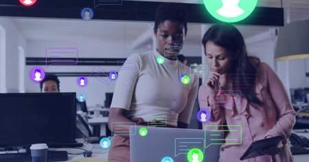 Collaborating coworkers examining data on laptop at open plan office desk, with floating icons. Professional, teamwork, modern, digital, communication, productivity, businessの写真素材