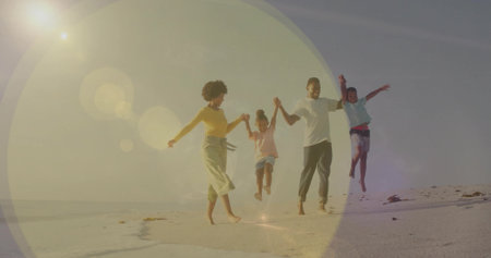 Jumping family of four wearing beachwear holding hands along sandy shoreline, with sun lens flare....