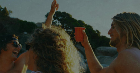 Gathering friends in swimwear raising red plastic cups on rocky beach cove, with baseball cap. Celebration, outdoor, leisure, camaraderie, vibrant, scenic, relaxedの写真素材