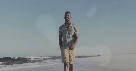 Strolling adult male wearing shirt and shorts admiring surf along sandy beach, with coastal scenery. Coastal, leisure, nature, serenity, outdoor, relaxed, vividの写真素材