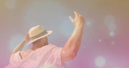 Man wearing fedora and pink off shoulder shirt lifting arms in studio, with pastel bokeh backdrop. Creative, fashion, aesthetic, confidence, leisure, artistic, vibrantの写真素材