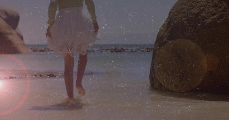 Child wearing light green top, tulle skirt wading into shallow water on sandy shore, with boulder. Playfulness, innocence, serenity, outdoor, leisure, summer, carefreeの写真素材