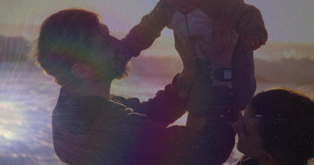 Hoisting father raising infant boy as crouching mother watching on beach at sunset, with lens flare. Family, beach, sunset, silhouette, togetherness, outdoor, leisureの写真素材