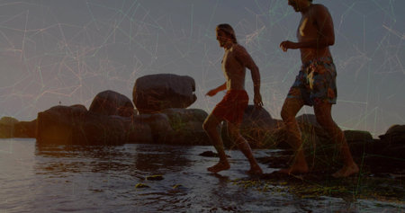 Walking shirtless men balancing across shallow water on beach at sunset, with colorful swim shorts....