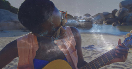 Woman strumming acoustic guitar wearing sunglasses, sleeveless shirt over swim top on sandy cove....