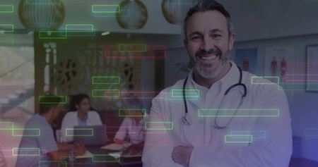 Smiling mature male doctor wearing lab coat and stethoscope in meeting room, with laptop and charts....