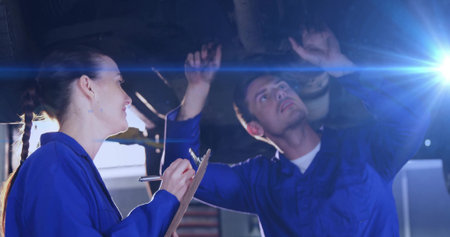 Two mechanics in coveralls inspecting undercarriage on vehicle lift at workshop, with clipboard pen....