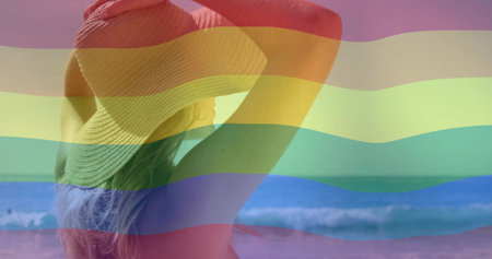 Facing waves, woman wearing swimsuit holding brim of sunhat on sandy beach, with rainbow overlay. Seascape, leisure, outdoor, nature, tranquility, carefree, lifestyleの写真素材