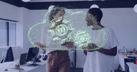 Reviewing cloud hologram with gears, two colleagues collaborating in modern office, with devices....