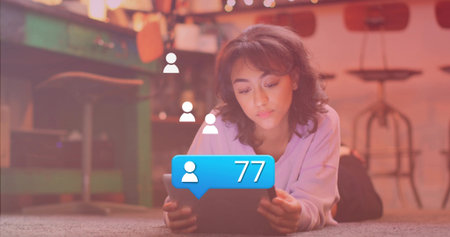 Lying woman watching tablet in carpeted living room, with floating icons and blue 77 bubble. Relaxation, digital interaction, warm lighting, cozy environment, social media, indoor leisure, casual lifestyleの写真素材