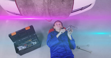 Female mechanic in coveralls lying under car using wrench and extension bar in garage. Industrial, repair, technical, equipment, professional, workshop, functionalの写真素材