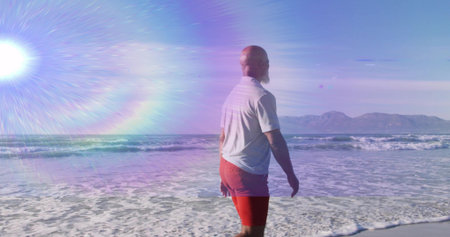 Gazing man wearing white T-shirt red trunks wading beach surf, with sun flare mountains, copy space....