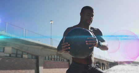 Shirtless man performing twisting exercise with black ball and smartwatch on beach walkway. Fitness,...