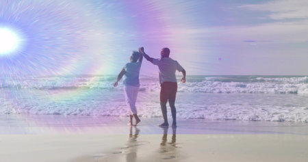 Dancing senior couple barefoot holding hands on beach shore, with breaking ocean waves, lens flare. Romantic, leisure, tranquility, coastal, serenity, vitality, sunsetの写真素材