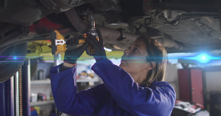 Working mechanic in blue coveralls using impact wrench under vehicle on hydraulic lift in workshop. Auto repair, industrial, workshop, tool, maintenance, professionalism, machineryの写真素材