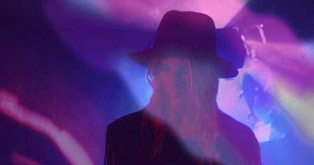 Standing adult woman wearing wide hat performing on nightclub stage, with colorful lights and haze....