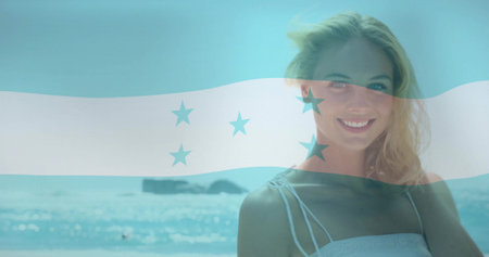 Smiling woman wearing white top standing on beach with flag overlay, waves and islets, copy space. Coastal, leisure, serene, vibrant, nature, lifestyle, outdoorsの写真素材