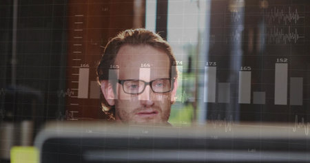 Man in eyeglasses analyzing data on monitor at office desk, with bar and line graphs overlay. Professional, technology, analytics, modern, workplace, business, digitalの写真素材