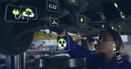Female mechanic in jumpsuit draining oil, inspecting car underside in garage, with lift, eco icons. Automotive, repair, sustainability, environmental, industrial, technology, workspaceの写真素材